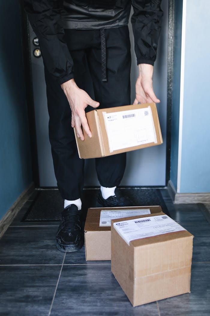 Person in dark clothing holding cardboard box in doorway with packages on the floor.